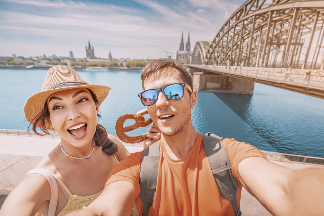 Date-Ideen in Köln: Ein junges Paar macht ein Selfie mit einer traditionellen deutschen Brezel vor dem Hintergrund des Kölner Doms und des Rheins.