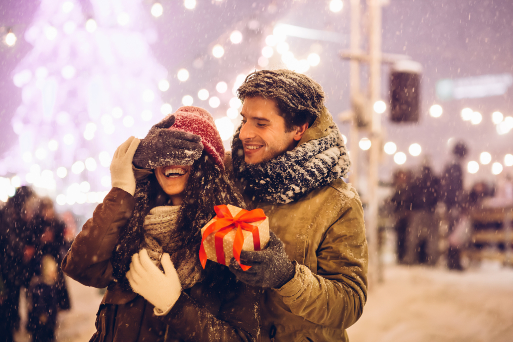 Winter Date-Ideen: Paar steht gemeinsam in der Stadt, während Freund Augen der Freundin schließt und Geschenk überreicht